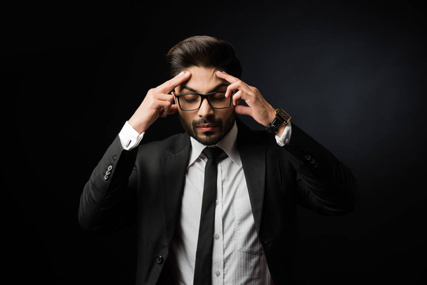 Stresszes indiai üzletembernek fejfájása vagy migrénes fájdalma van. Álló elszigetelt felett fekete háttér visel szemüveg - Fotó, kép