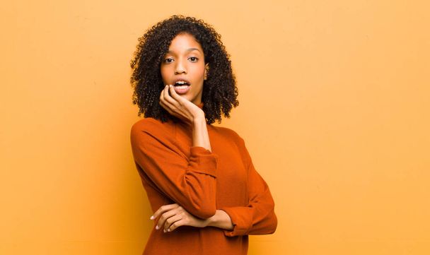 young pretty black woman open-mouthed in shock and disbelief, with hand on cheek and arm crossed, feeling stupefied and amazed against orange wall - Fotó, kép