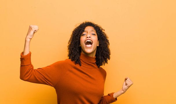fiatal csinos fekete nő kiabál diadalmasan, úgy néz ki, mint izgatott, boldog és meglepett győztes, ünneplő narancssárga fal - Fotó, kép