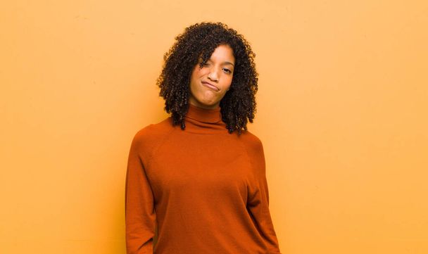 young pretty black woman looking happy and friendly, smiling and winking an eye at you with a positive attitude against orange wall - Foto, immagini