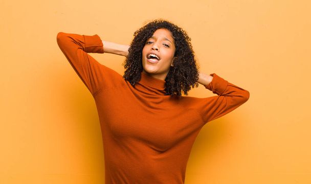 young pretty black woman smiling and feeling relaxed, satisfied and carefree, laughing positively and chilling against orange wall - Foto, afbeelding