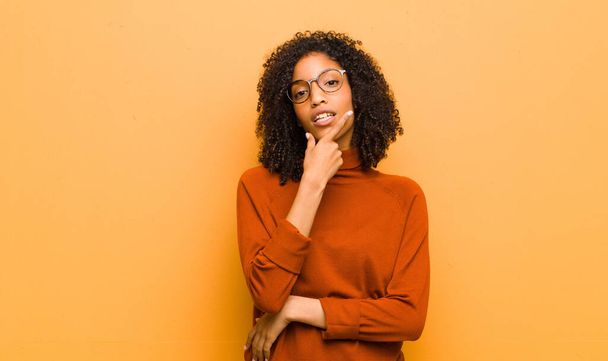 young pretty black woman looking serious, thoughtful and distrustful, with one arm crossed and hand on chin, weighting options against orange wall - Foto, immagini