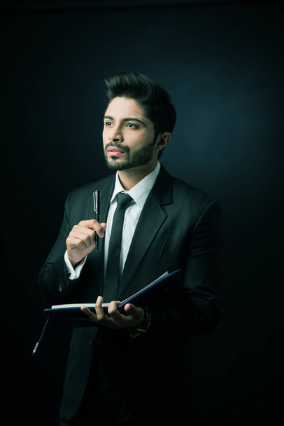 Hombre de negocios indio escribiendo en un diario mientras está de pie sobre fondo negro, iluminación malhumorada, enfoque selectivo
 - Foto, imagen