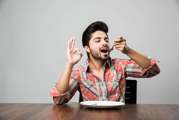 empty plate and Indian man with beard holding spoon and fork, wearing checkered shirt and sitting at table  - 写真・画像