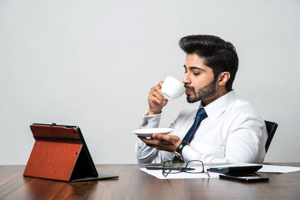 Indian businessman having coffee / tea at work / office, sitting at desk or table holding cup and saucer  - Photo, Image