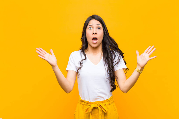 young pretty latin woman looking shocked and astonished, with jaw dropped in surprise when realizing something unbelievable against orange wall - 写真・画像