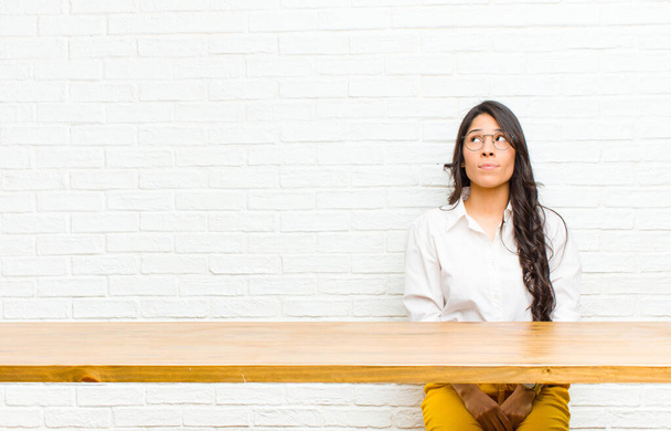young  pretty latin woman with a worried, confused, clueless expression, looking up to copy space, doubting sitting in front of a table - Photo, image