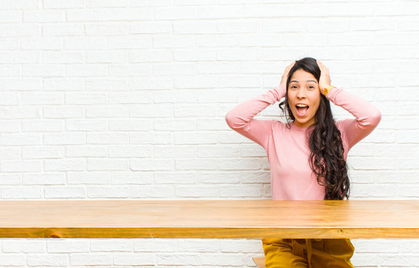 young  pretty latin woman raising hands to head, open-mouthed, feeling extremely lucky, surprised, excited and happy sitting in front of a table - 写真・画像
