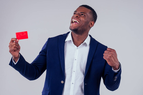 stylish african american in costume black pants and jacket with a big shirt collar holding red credit plastic card in studio on white background, badge my name is - Φωτογραφία, εικόνα