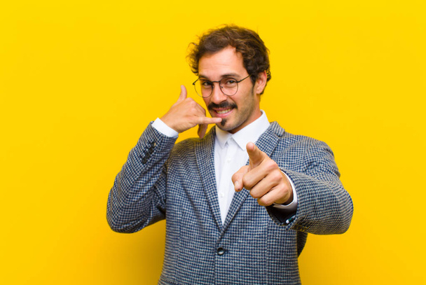 young handsome man smiling cheerfully and pointing to camera while making a call you later gesture, talking on phone against orange wall - Foto, imagen