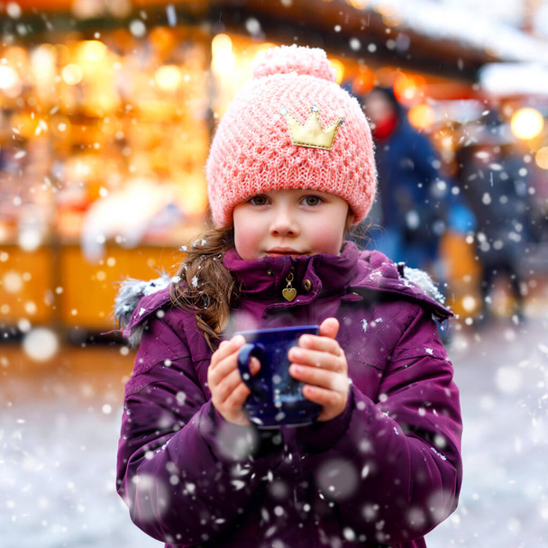 kleines süßes Mädchen mit einer Tasse dampfender heißer Schokolade oder Kinderpunsch. Glückskind auf dem Weihnachtsmarkt in Deutschland. Traditionelle Freizeit für Familien an Weihnachten. - Foto, Bild