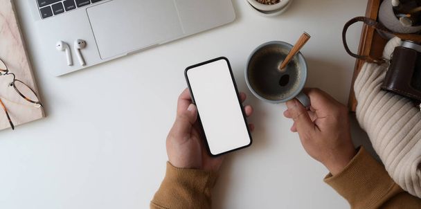 Visão superior do homem olhando para o smartphone tela em branco enquanto segurando xícara de café no local de trabalho acolhedor
 - Foto, Imagem