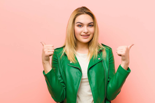 young pretty blonde woman smiling joyfully and looking happy, feeling carefree and positive with both thumbs up against pink flat wall - Fotoğraf, Görsel