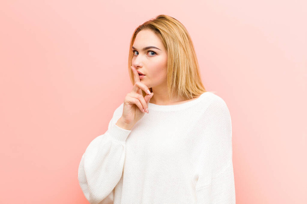 young pretty blonde woman asking for silence and quiet, gesturing with finger in front of mouth, saying shh or keeping a secret against pink flat wall - Фото, изображение