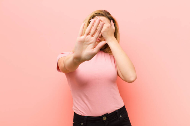 young pretty blonde woman covering face with hand and putting other hand up front to stop camera, refusing photos or pictures against pink flat wall - Foto, afbeelding