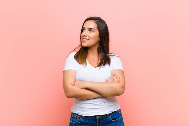 young pretty hispanic woman feeling happy, proud and hopeful, wondering or thinking, looking up to copy space with crossed arms against pink wall - Фото, изображение