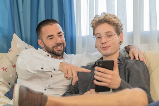 Ein bärtiger Mann zeigt auf ein Foto auf dem Smartphone seiner Freunde mit blonden Haaren und Brille, die beide entspannt auf dem Sofa sitzen. Konzept der Koexistenz und Geselligkeit. - Foto, Bild