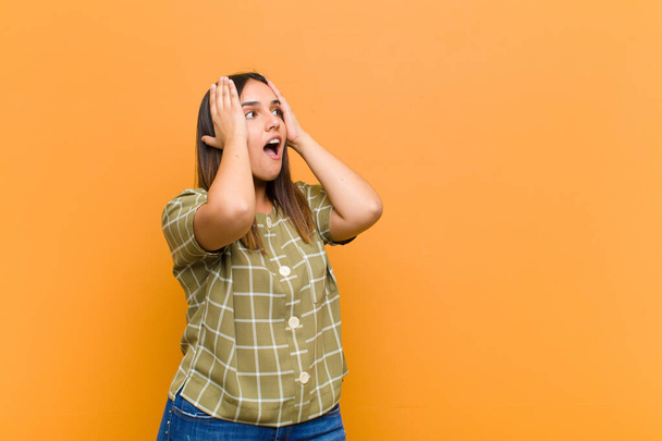 young pretty woman with open mouth, looking horrified and shocked because of a terrible mistake, raising hands to head isolated against orange wall - Foto, imagen
