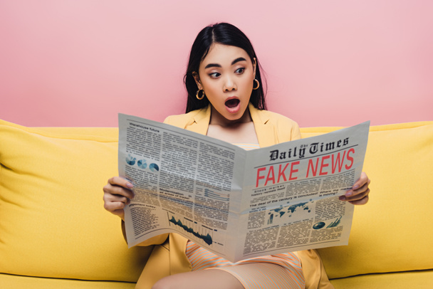 shocked asian woman holding newspaper with fake news isolated on pink - 写真・画像