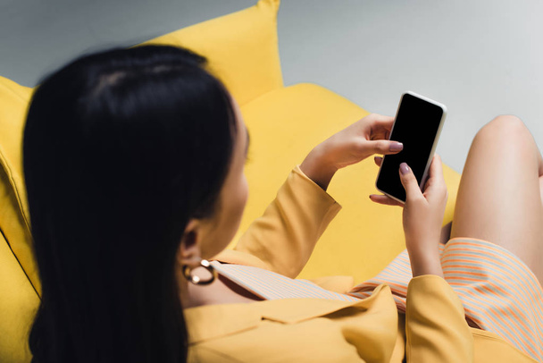 overhead view of asian woman holding smartphone while sitting on yellow couch isolated on grey - Фото, зображення
