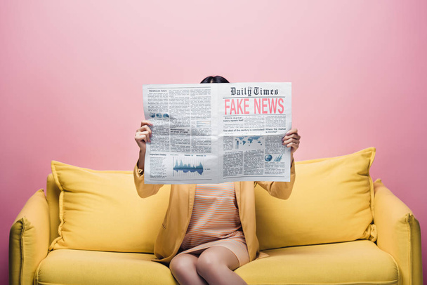 asian woman holding newspaper with fake news in front of face while sitting on yellow sofa isolated on pink - Photo, Image