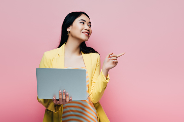 happy asian woman in yellow outfit holding laptop and pointing with finger away isolated on pink - Photo, Image