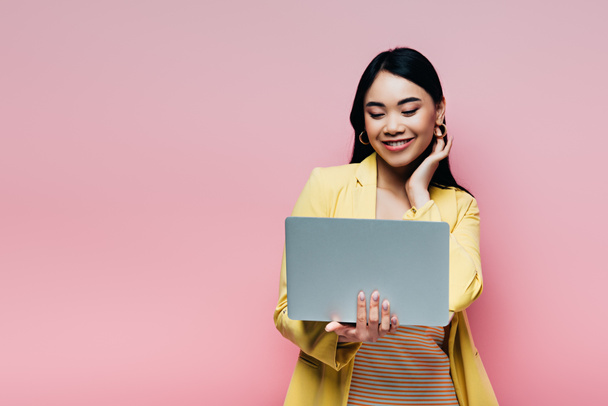 happy asian woman in yellow outfit holding laptop isolated on pink - Foto, Imagen