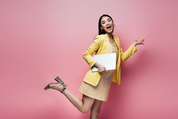 excited happy asian woman in yellow outfit posing with laptop isolated on pink - Foto, Imagem