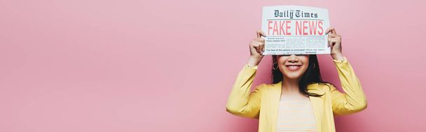 panoramic shot of happy asian woman in yellow outfit holding newspaper with fake news isolated on pink - Foto, Bild