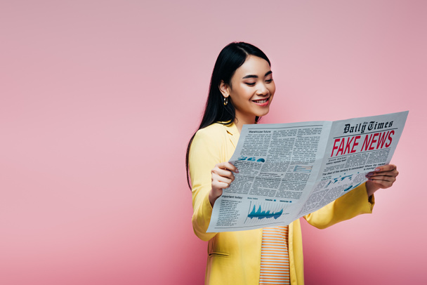 счастливая азиатка в желтом наряде читает газету с фальшивыми новостями изолированы на розовый
 - Фото, изображение