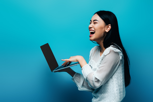 happy asian woman in white blouse holding laptop and laughing on blue background - Foto, immagini