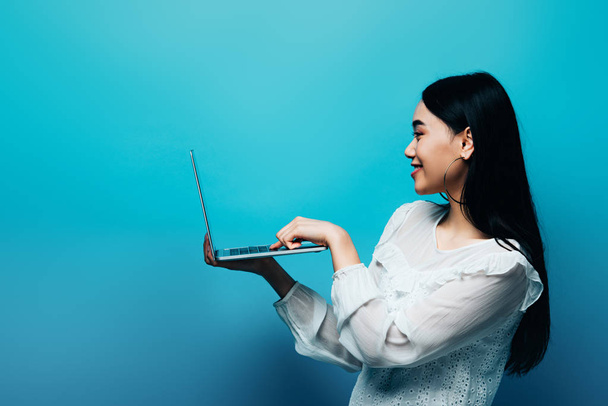 side view of smiling asian woman in white blouse holding laptop on blue background - Foto, immagini