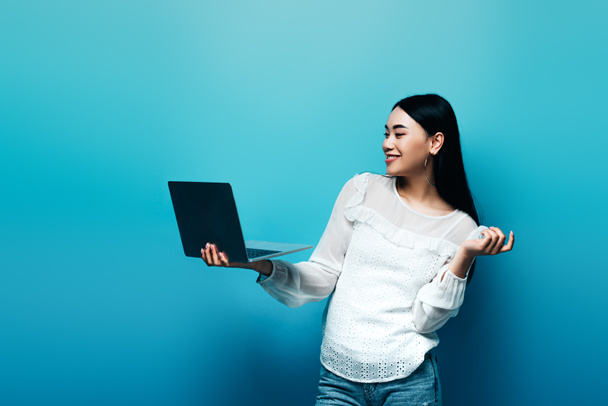 smiling asian woman in white blouse holding laptop on blue background - Valokuva, kuva