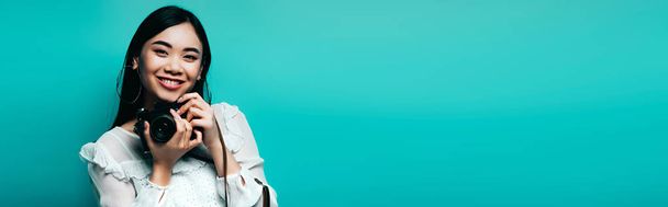 happy asian woman in white blouse holding digital camera on blue background, panoramic shot - Foto, Bild