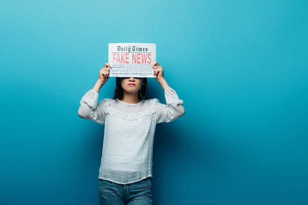 Asiatin in weißer Bluse hält Zeitung mit Fake News auf blauem Hintergrund - Foto, Bild