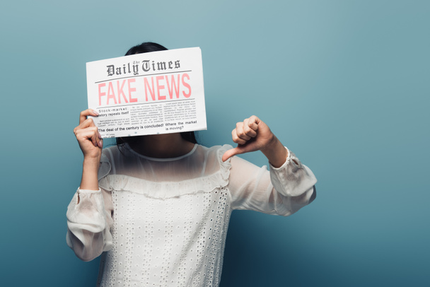woman in white blouse holding newspaper with fake news and showing thumb down on blue background - Fotografie, Obrázek