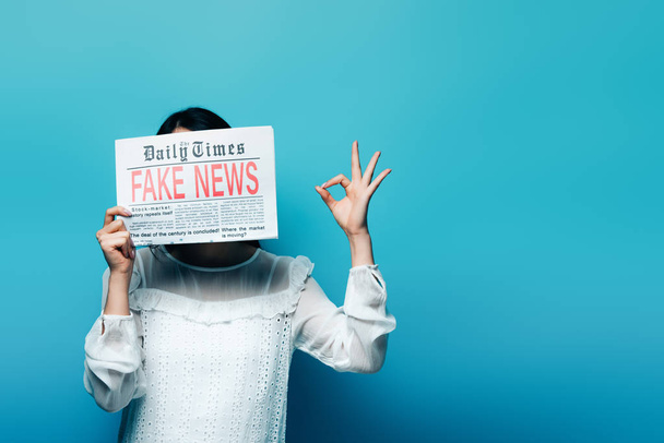 Beyaz bluzlu kadın elinde sahte haberlerle gazete tutuyor ve mavi arka planda "Tamam" işareti gösteriyor. - Fotoğraf, Görsel