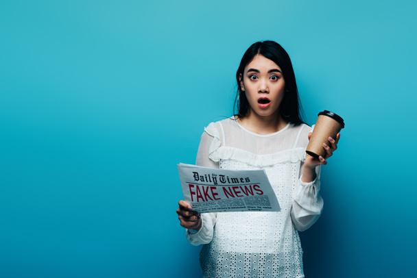 шокированная азиатка в белой блузке, держащая газету с фальшивыми новостями и бумажной чашкой на синем фоне
 - Фото, изображение