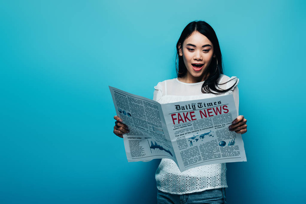 shocked asian woman in white blouse reading newspaper with fake news on blue background - Fotoğraf, Görsel