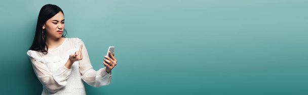 tiro panorámico de mujer asiática insatisfecha en blusa blanca mostrando el dedo medio en la pantalla del teléfono inteligente sobre fondo verde
 - Foto, imagen