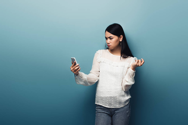 mujer asiática insatisfecha en blusa blanca sosteniendo teléfono inteligente sobre fondo azul
 - Foto, imagen