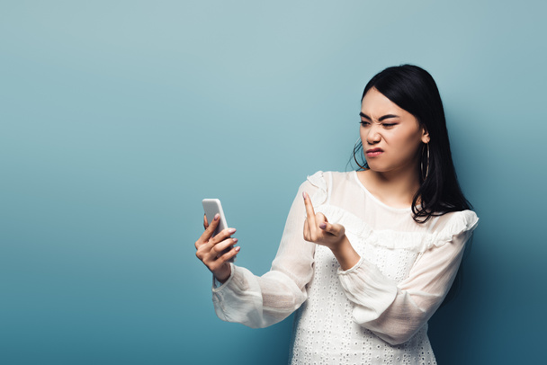 dissatisfied asian woman in white blouse showing middle finger in smartphone screen on blue background - Photo, Image