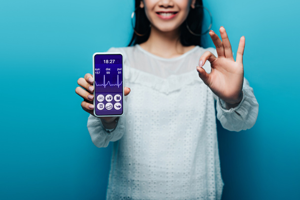 oříznutý pohled na usmívající se asijské ženy v bílé blůze ukazující ok znamení a smartphone s aplikací zdravotní péče na modrém pozadí - Fotografie, Obrázek