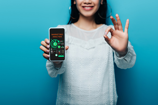 обрезанный вид улыбающейся азиатской женщины в белой блузке показывает знак ОК и смартфон с приложением маркетингового анализа на синем фоне
 - Фото, изображение