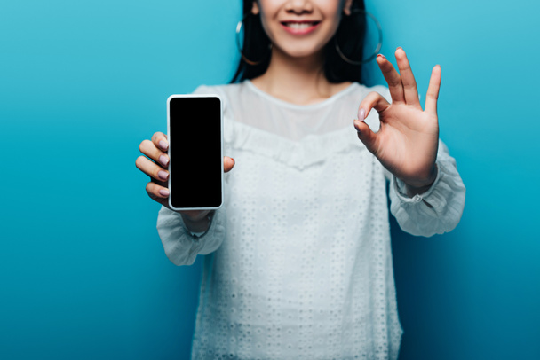 обрезанный вид улыбающейся азиатской женщины в белой блузке показывает знак ОК и смартфон с чистым экраном на синем фоне
 - Фото, изображение