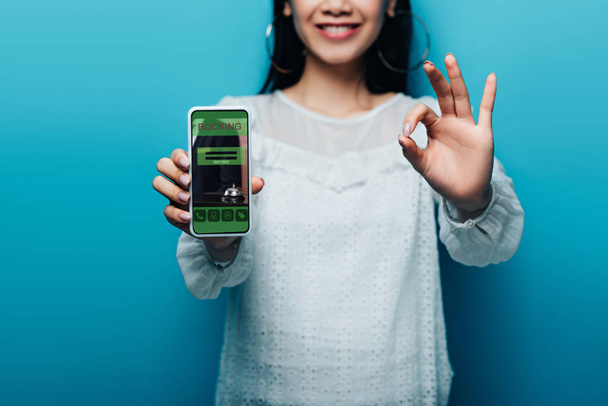 cropped view of smiling asian woman in white blouse showing ok sign and smartphone with booking app on blue background - 写真・画像