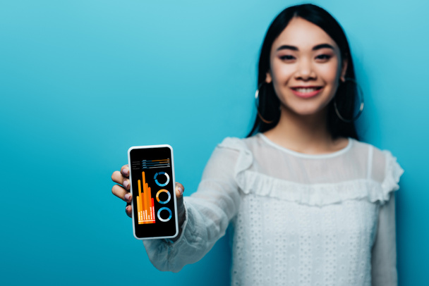 selective focus of smiling asian woman in white blouse holding smartphone with business app on blue background - Foto, imagen
