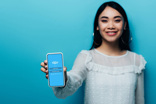 kyiv, ukraine - 15. Juli 2019: selektiver Fokus einer lächelnden asiatischen Frau in weißer Bluse mit Smartphone mit Skype-App auf blauem Hintergrund - Foto, Bild