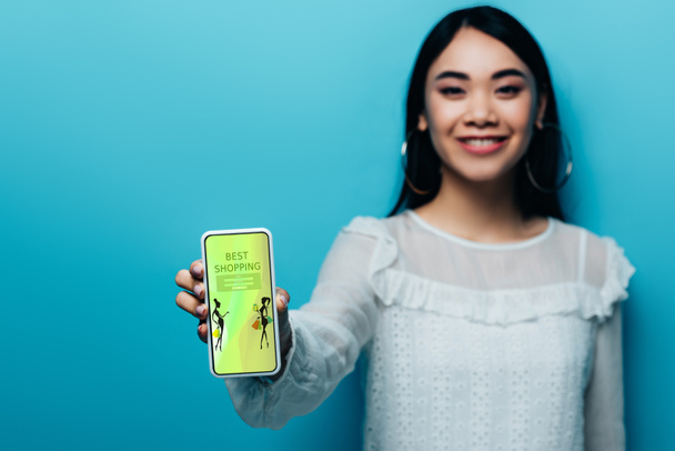 Selektiver Fokus einer lächelnden asiatischen Frau in weißer Bluse mit Smartphone und Mode-Shopping-App auf blauem Hintergrund - Foto, Bild