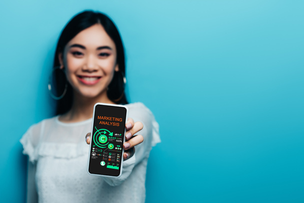 селективный фокус улыбающейся азиатской женщины в белой блузке, держащей смартфон с приложением маркетингового анализа на синем фоне
 - Фото, изображение